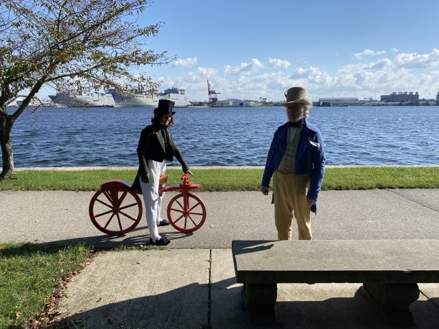 two men in 1820s clothing stand in front of Baltimore harbor. They are wearing tall hats and long coats. The man on the left stands astride a red velocipede.